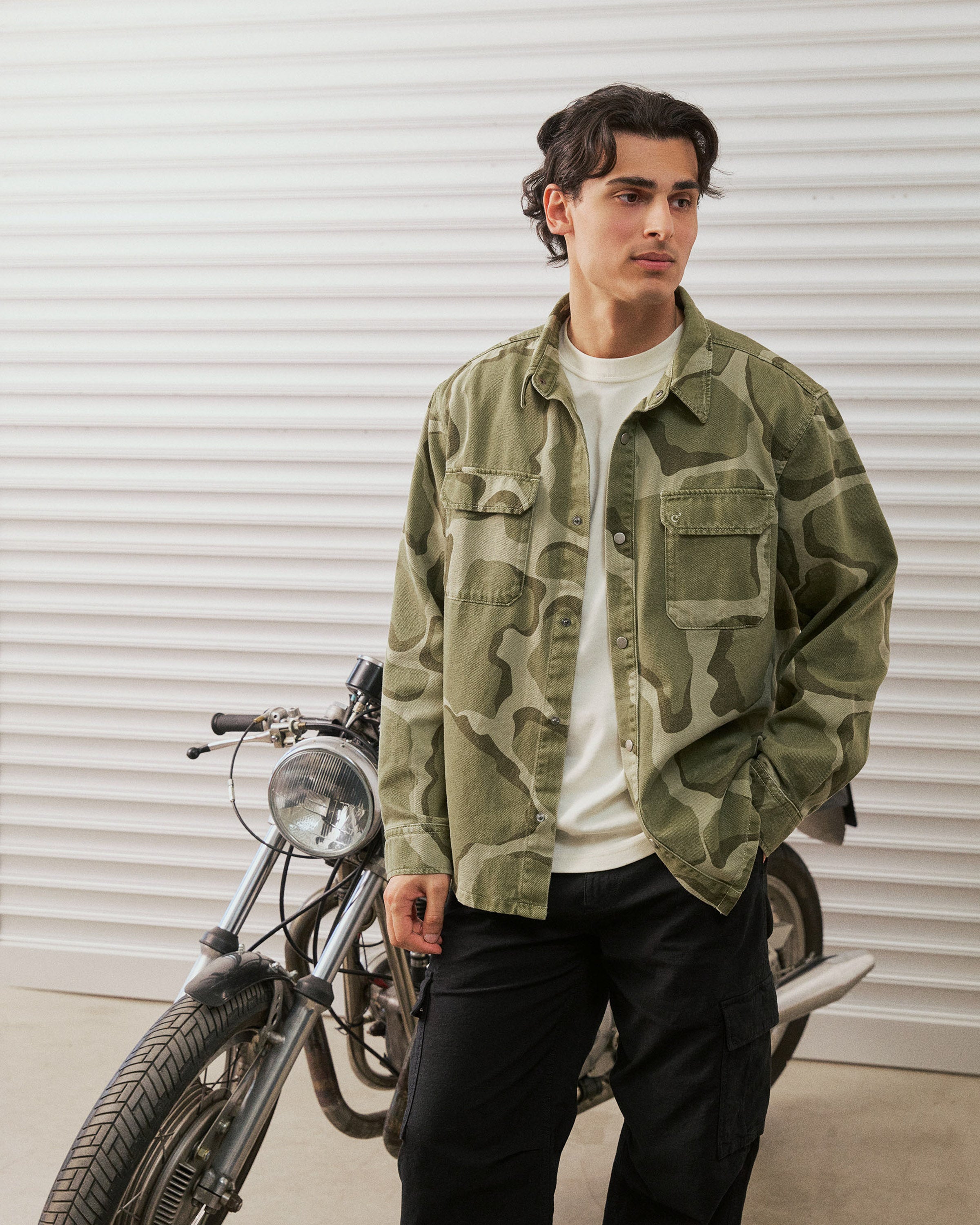 The Bilbao Overshirt in camo — worn while standing next to a vintage motorcycle