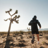 A man running in a desert landscape with Joshua trees, viewed from behind. He is wearing The Running Jacket in black, which features "ATHLÉTIQUE" and the c'est normal logo on the back.