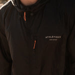 A close-up shot of a person wearing The Running Jacket in black. The image highlights the "ATHLÉTIQUE c'est normal" reflective branding on the chest, along with the contrast orange zipper pullers on the hood and front pocket.