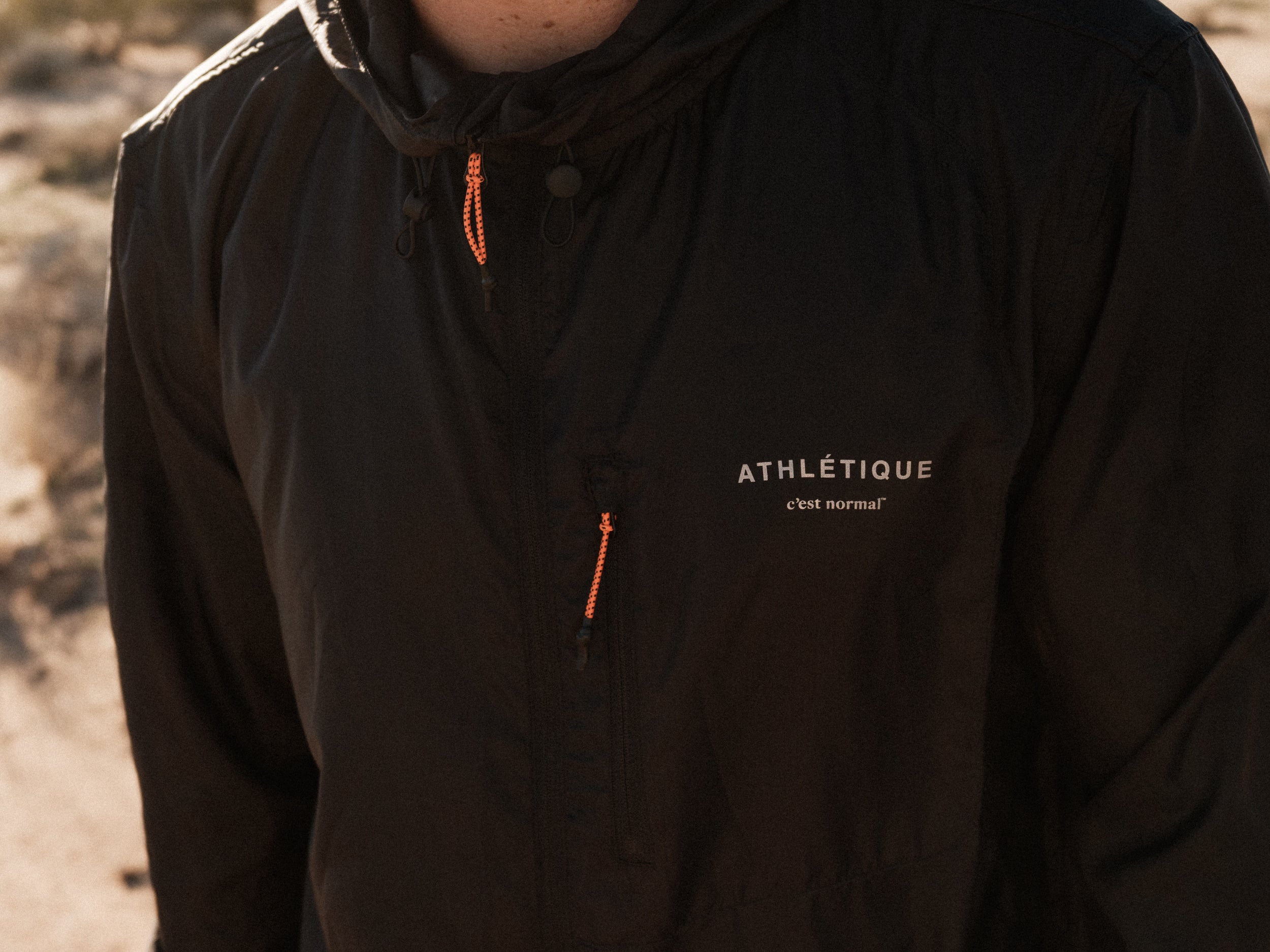 A close-up shot of a person wearing The Running Jacket in black. The image highlights the "ATHLÉTIQUE c'est normal" reflective branding on the chest, along with the contrast orange zipper pullers on the hood and front pocket.