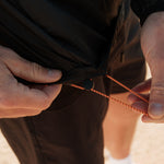 A close-up shot of a person adjusting the orange and black drawstring on the hem of The Running Jacket in black.