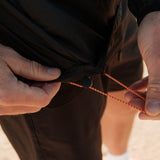 A close-up shot of a person adjusting the orange and black drawstring on the hem of The Running Jacket in black.