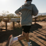 A person running on a dirt path wearing The Running Jacket in Sage Green, viewed from behind. The image captures the motion of the runner in a desert landscape.