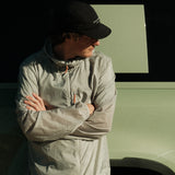 A man wearing The Running Jacket in sage green, with his arms crossed, standing next to a vehicle. He is also wearing a black c'est normal cap and looking down and to the right.