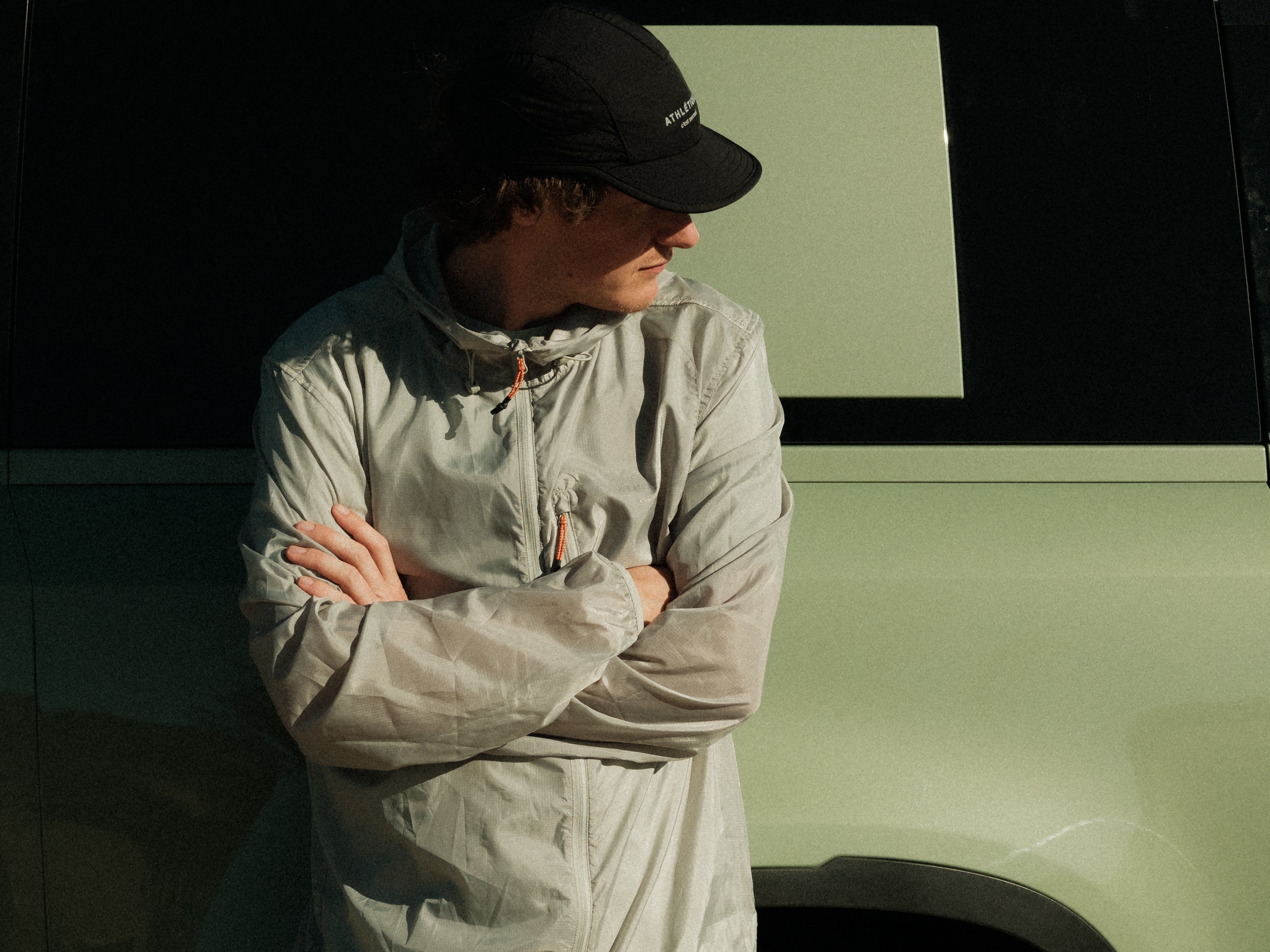 A man wearing The Running Jacket in sage green, with his arms crossed, standing next to a vehicle. He is also wearing a black c'est normal cap and looking down and to the right.