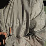 A close-up shot of a person wearing The Running Jacket in Sage Green, adjusting a black sports watch. The image highlights the jacket's lightweight fabric, contrast zipper pullers, and the reflective 'ATHLÉTIQUE c'est normal' logo on the chest.