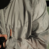 A close-up shot of a person wearing The Running Jacket in Sage Green, adjusting a black sports watch. The image highlights the jacket's lightweight fabric, contrast zipper pullers, and the reflective 'ATHLÉTIQUE c'est normal' logo on the chest.