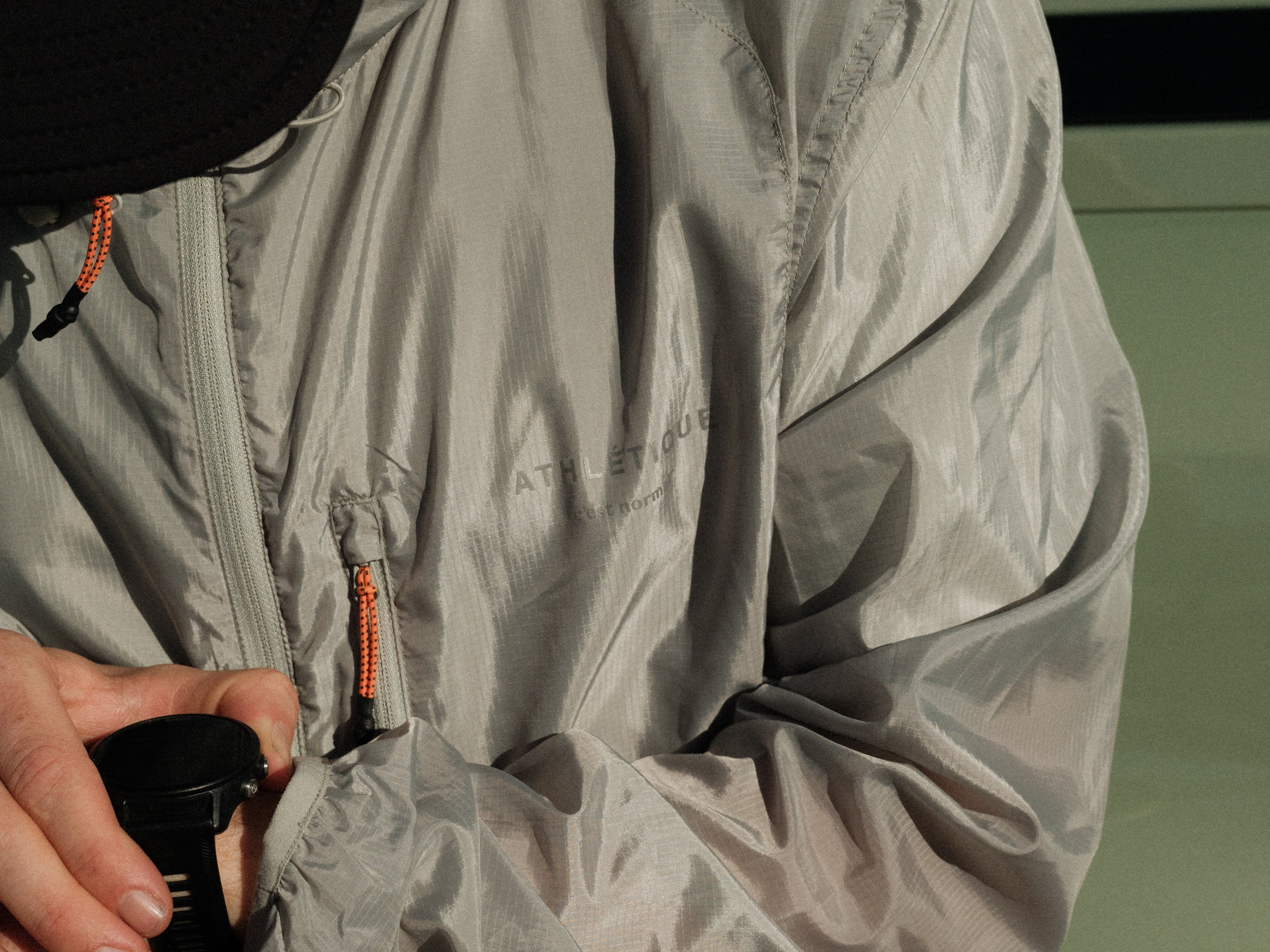 A close-up shot of a person wearing The Running Jacket in Sage Green, adjusting a black sports watch. The image highlights the jacket's lightweight fabric, contrast zipper pullers, and the reflective 'ATHLÉTIQUE c'est normal' logo on the chest.