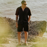 The Holiday Linen Short in black — worn with a matching linen shirt by the sea