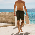 The Board Shorts in khaki — back view as a man walks toward the ocean holding a towel