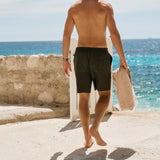 The Board Shorts in khaki — back view as a man walks toward the ocean holding a towel