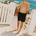 The Board Shorts in black — worn by a man holding a beach towel by the ocean