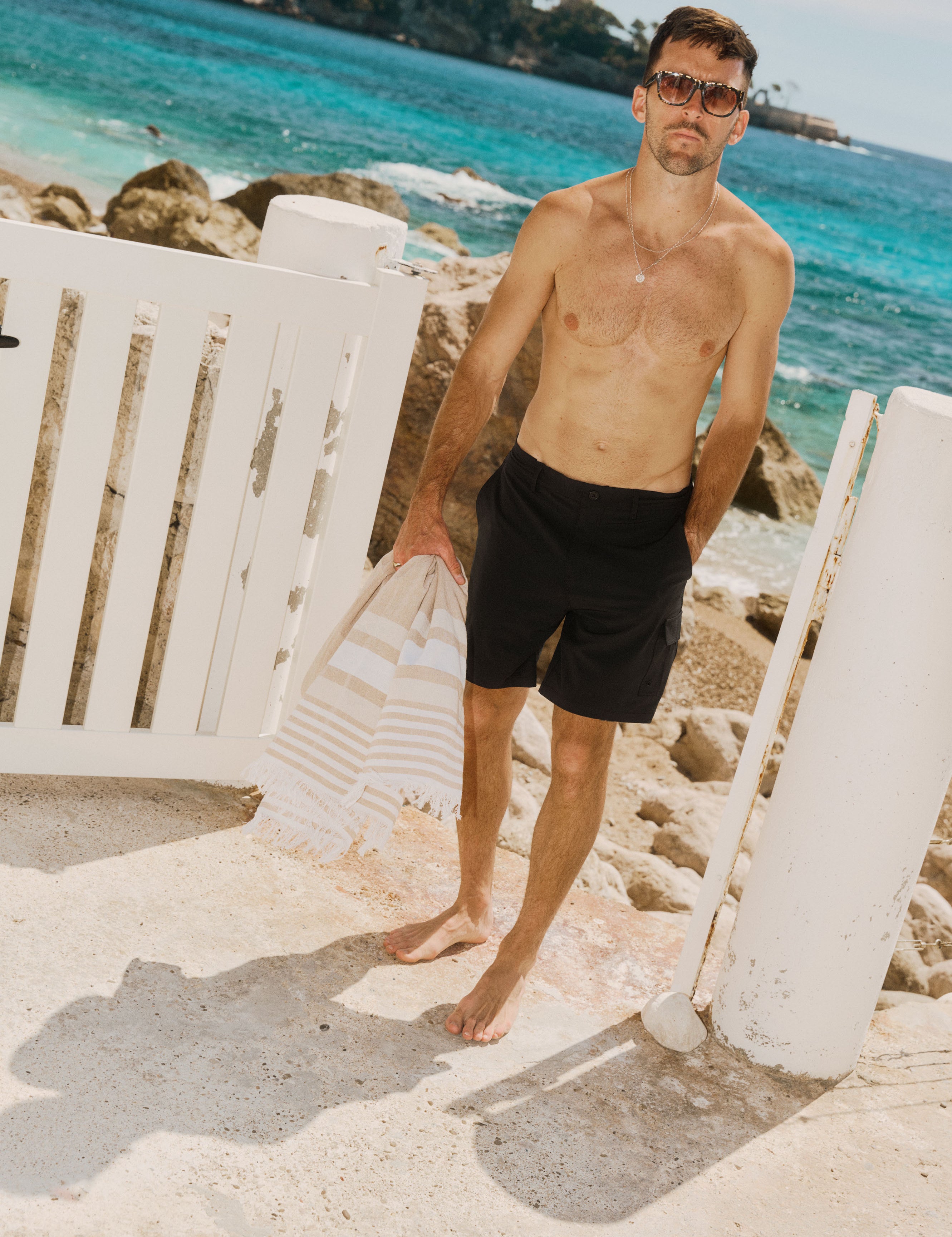 The Board Shorts in black — worn by a man holding a beach towel by the ocean