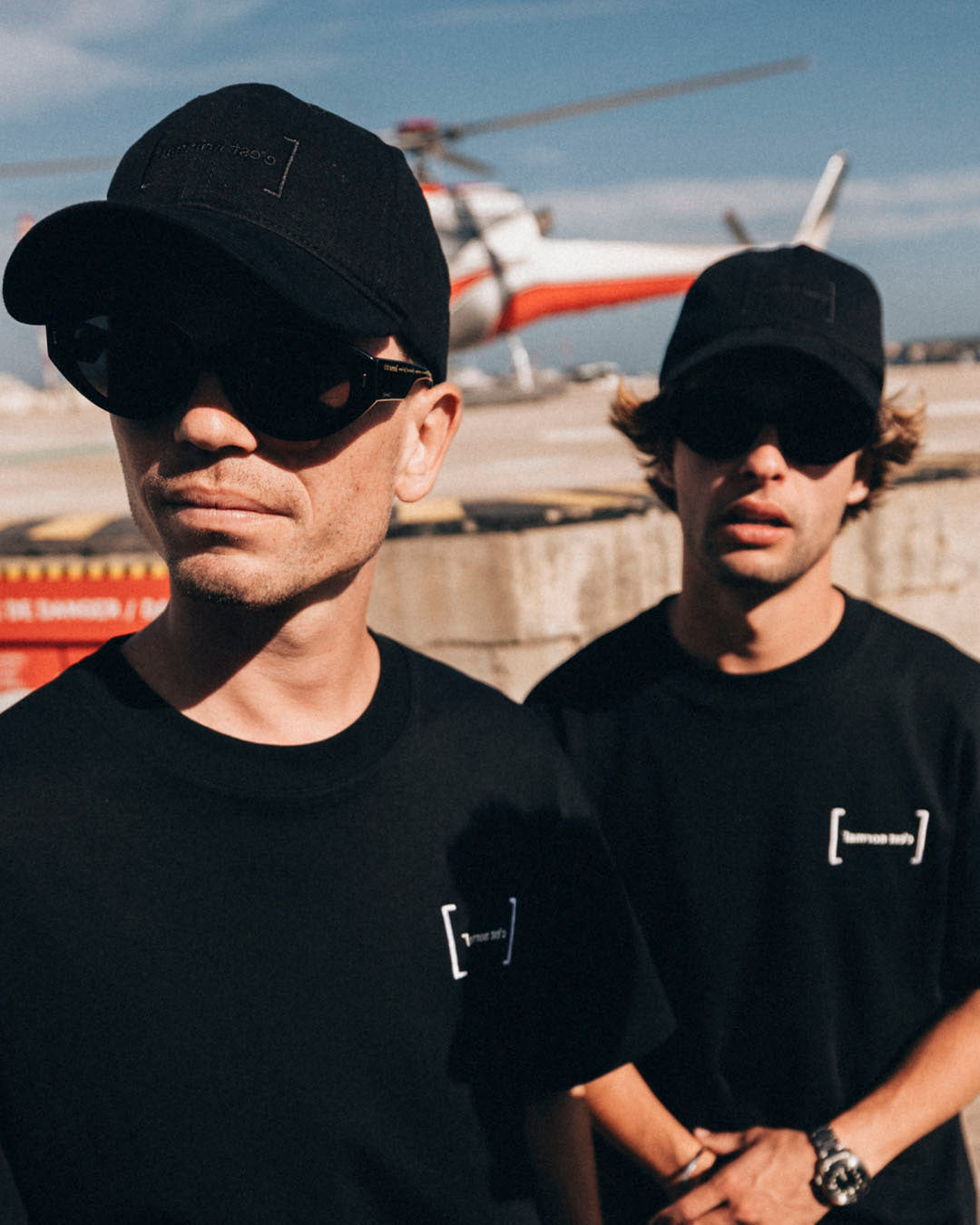 La Cap in black — worn by two men on an airfield with a helicopter