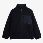 A front view of The Reversible Pile Jacket in black, laid flat on a white background. The image showcases the cozy pile fleece side, featuring a zippered chest pocket with an orange c'est normal logo, two zip hand pockets, and cinch cords at the hem.