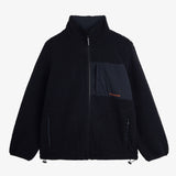 A front view of The Reversible Pile Jacket in black, laid flat on a white background. The image showcases the cozy pile fleece side, featuring a zippered chest pocket with an orange c'est normal logo, two zip hand pockets, and cinch cords at the hem.
