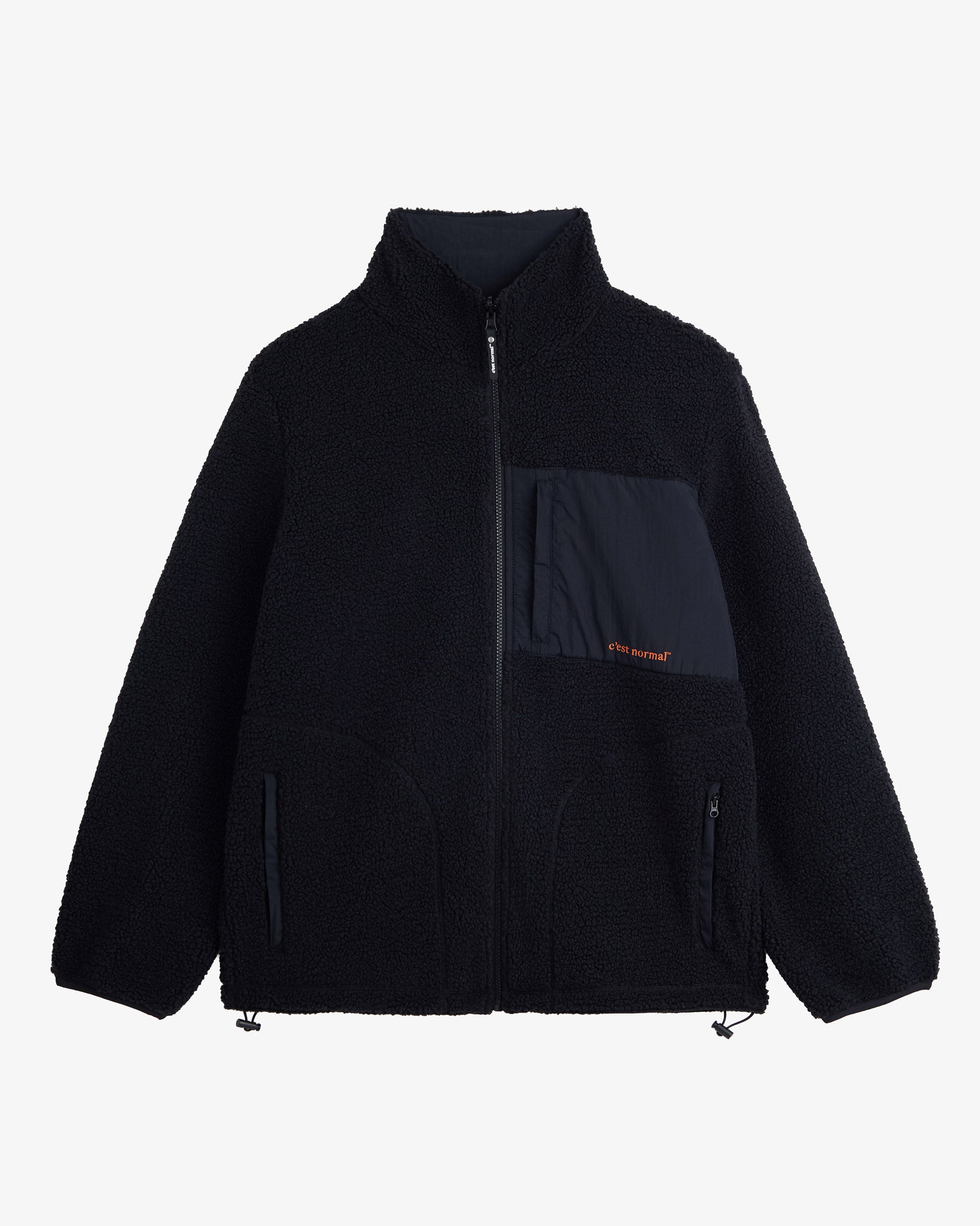 A front view of The Reversible Pile Jacket in black, laid flat on a white background. The image showcases the cozy pile fleece side, featuring a zippered chest pocket with an orange c'est normal logo, two zip hand pockets, and cinch cords at the hem.