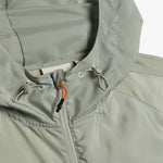 A close-up shot of the hood and neckline of The Running Jacket in Sage Green, highlighting the drawstring toggles and the contrast orange and black zipper puller. | c'est normal