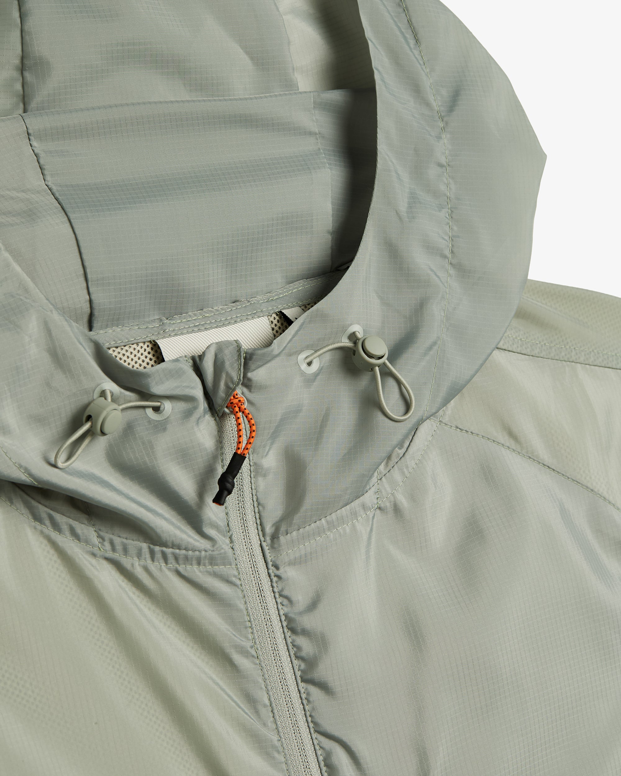 A close-up shot of the hood and neckline of The Running Jacket in Sage Green, highlighting the drawstring toggles and the contrast orange and black zipper puller. | c'est normal