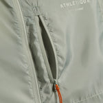 A detailed close-up of The Running Jacket in sage green, focusing on the front zippered pocket with an orange and black contrast zipper puller. The reflective ATHLÉTIQUE and c'est normal logos are printed above the pocket.