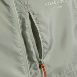A detailed close-up of The Running Jacket in sage green, focusing on the front zippered pocket with an orange and black contrast zipper puller. The reflective ATHLÉTIQUE and c'est normal logos are printed above the pocket.