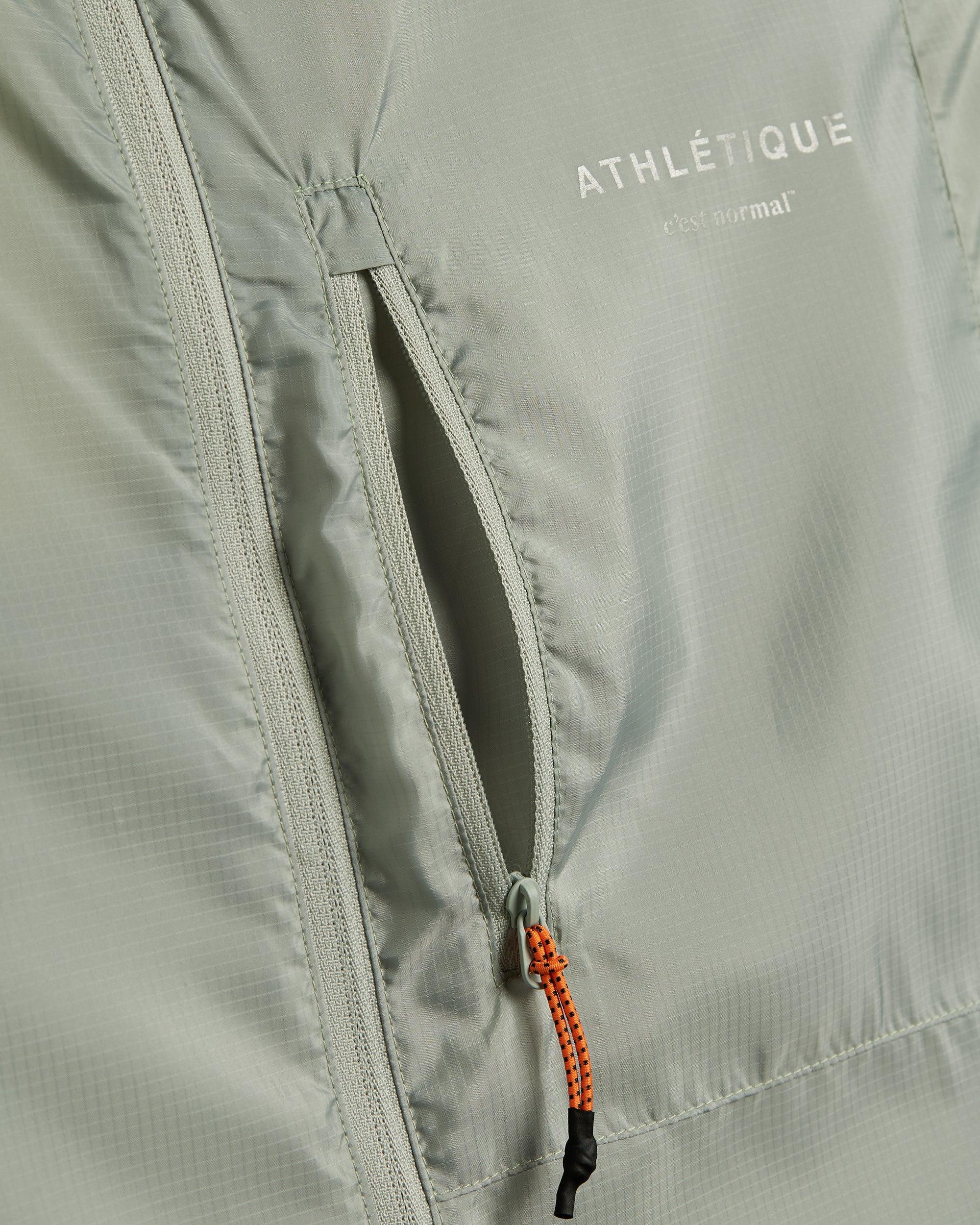 A detailed close-up of The Running Jacket in sage green, focusing on the front zippered pocket with an orange and black contrast zipper puller. The reflective ATHLÉTIQUE and c'est normal logos are printed above the pocket.