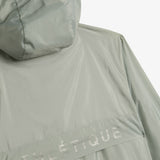 A close-up shot of the back of The Running Jacket in Sage Green, laid flat against a white background. The image focuses on the back ventilation panel, hood, and reflective ATHLÉTIQUE branding.