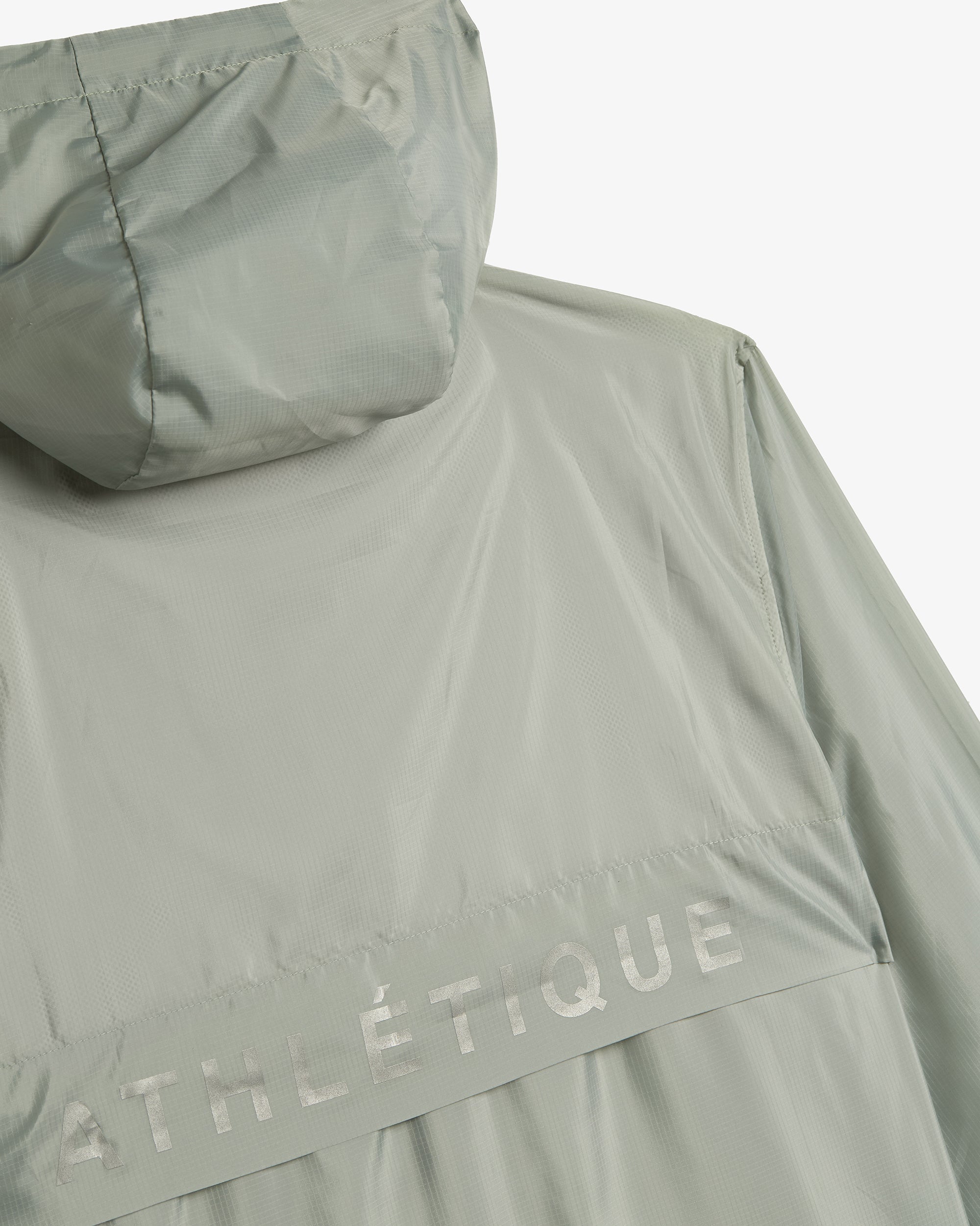 A close-up shot of the back of The Running Jacket in Sage Green, laid flat against a white background. The image focuses on the back ventilation panel, hood, and reflective ATHLÉTIQUE branding.