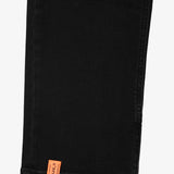 A detailed close-up of the ankle of The Jeans in black, highlighting the side zipper and the orange c'est normal tag at the hem.