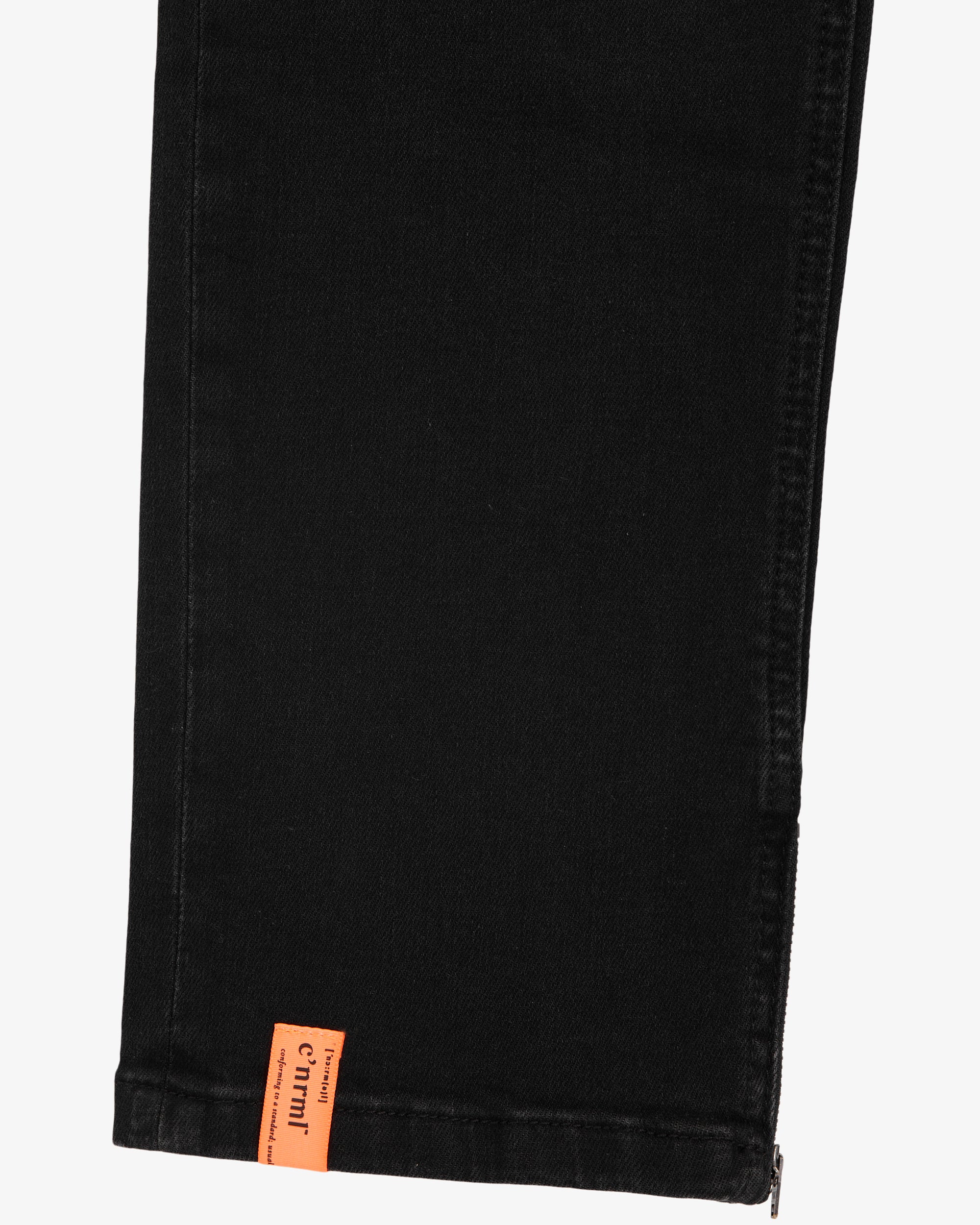 A detailed close-up of the ankle of The Jeans in black, highlighting the side zipper and the orange c'est normal tag at the hem.