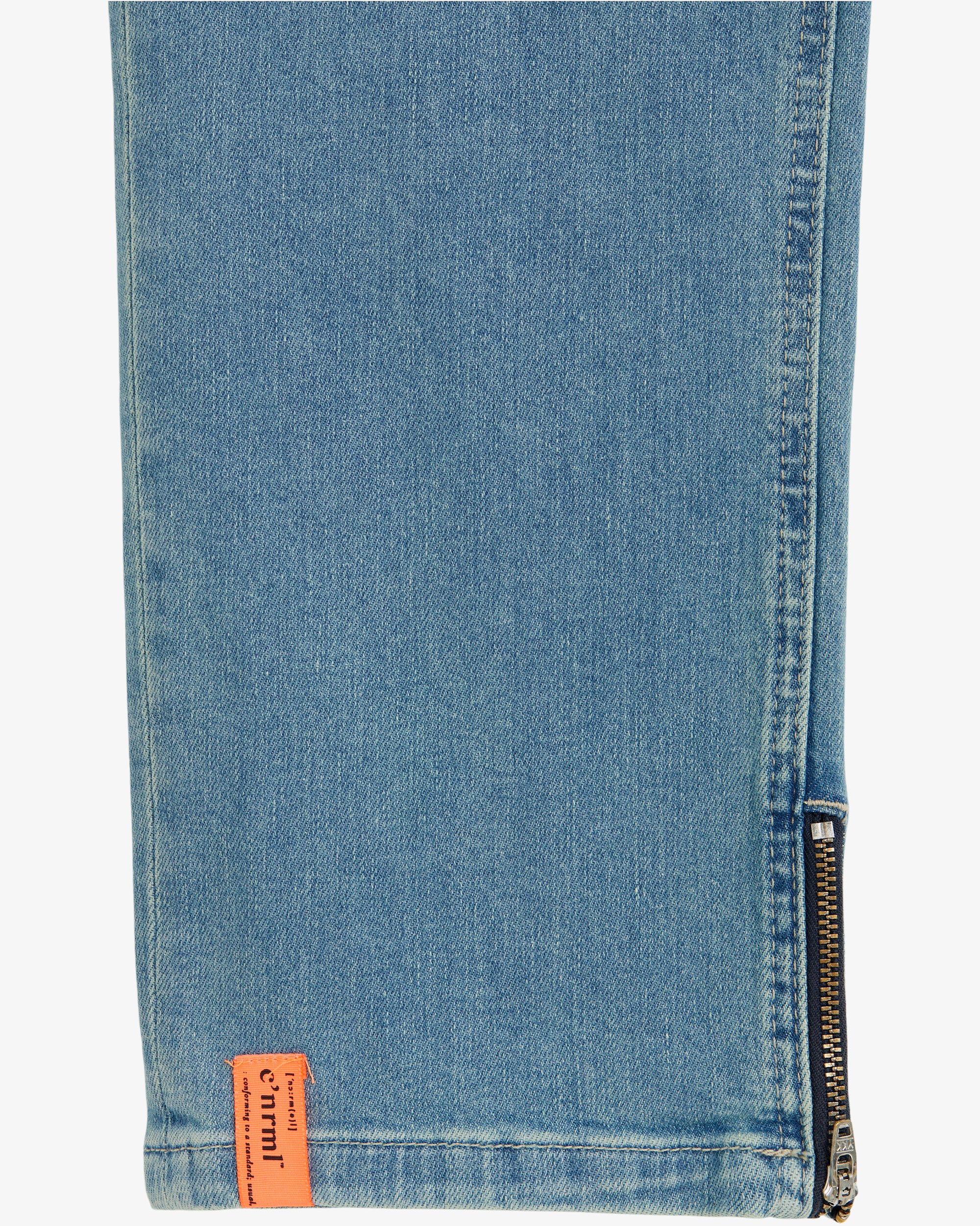 A close-up of the ankle hem of blue jeans, highlighting a metal side zipper and a small orange c'est normal tag.
