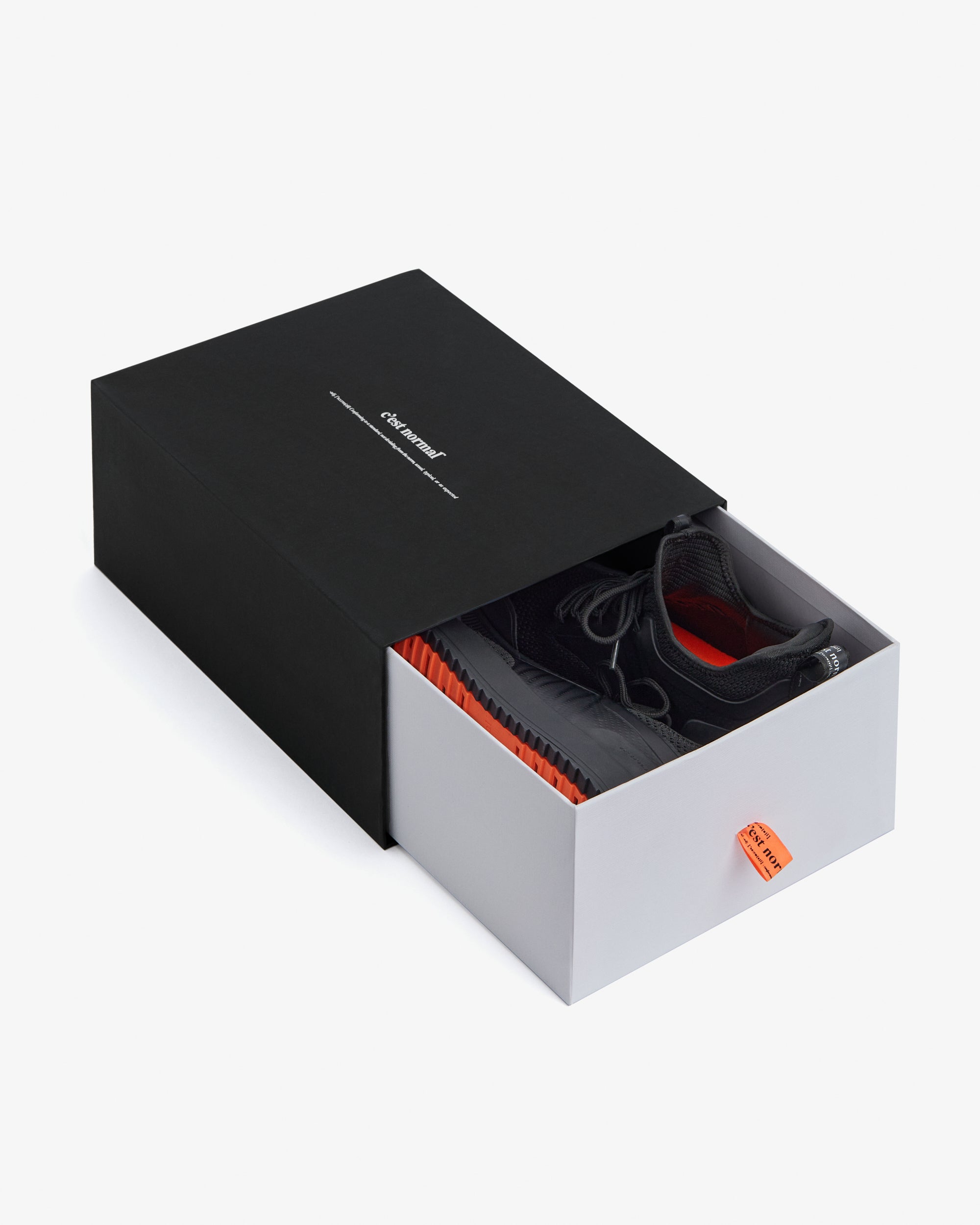 The Sneakers 2.0 in black, presented in a black and grey c'est normal shoebox with an orange pull tab on a white background.