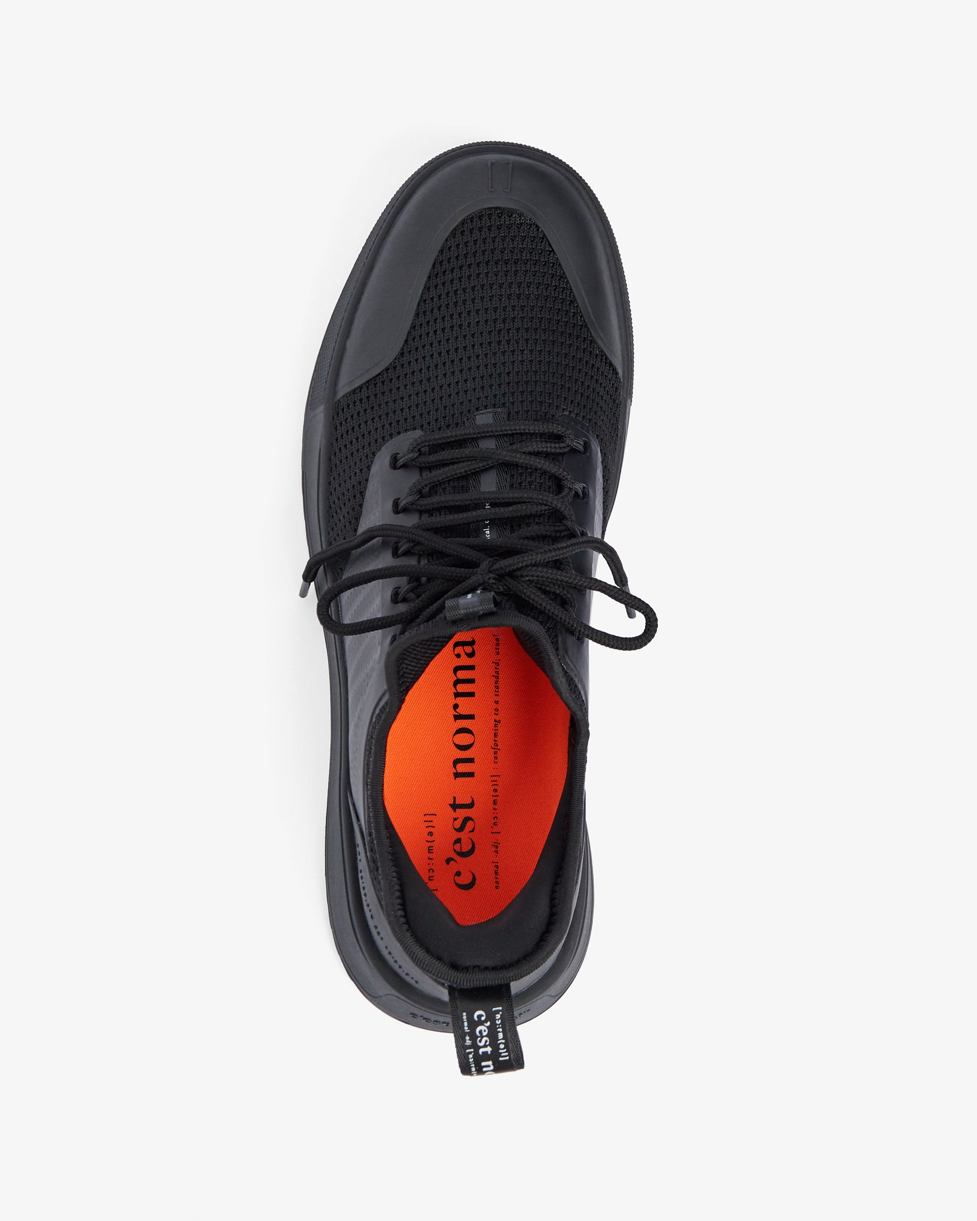 A top-down view of The Sneakers 2.0 in black, laid flat on a white background. The image highlights the tone on tone mesh upper, black laces, and the original orange insole featuring the c'est normal logo.