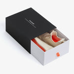 A pair of The Sneakers 2.0 in cream resting inside a partially opened black and white shoebox on a white background. The image highlights the cream mesh upper, the original orange rubber grid sole, and the c'est normal logo on the box.
