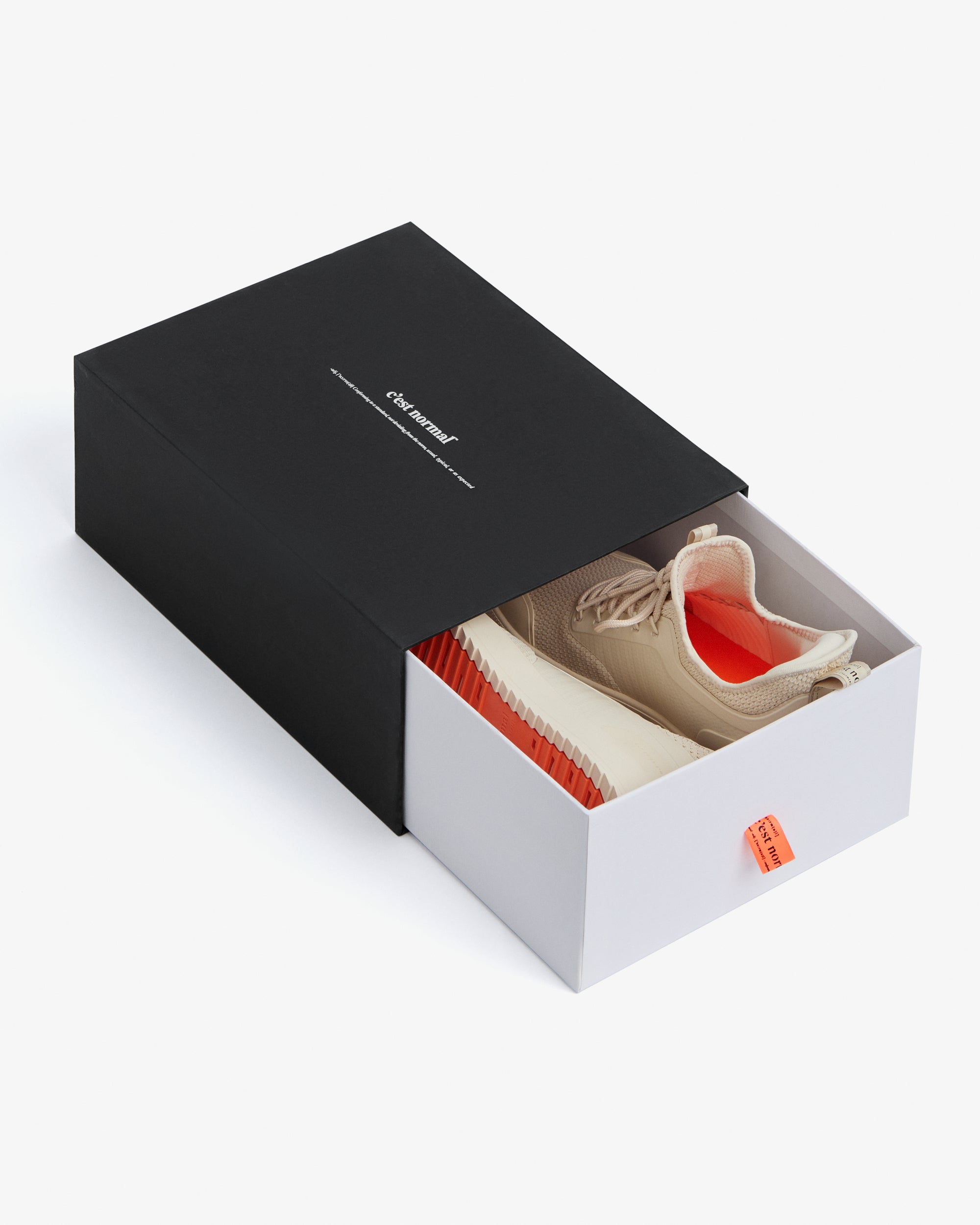 A pair of The Sneakers 2.0 in cream resting inside a partially opened black and white shoebox on a white background. The image highlights the cream mesh upper, the original orange rubber grid sole, and the c'est normal logo on the box.