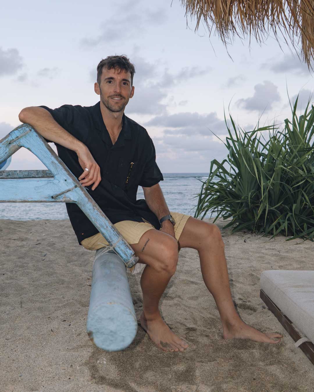 The Linen Resort Shirt in black — worn while sitting on a blue boat frame on a beach