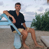The Linen Resort Shirt in black — worn while sitting on a blue boat frame on a beach