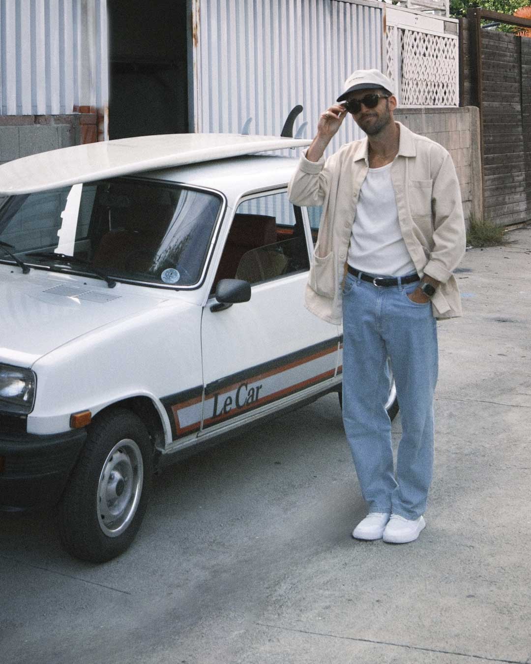 A man wearing The Workwear Overshirt in cream, a c'est normal cap, and sunglasses, stands next to a white vintage car with a surfboard on its roof. He is wearing the overshirt unbuttoned over a white t-shirt and light blue jeans.