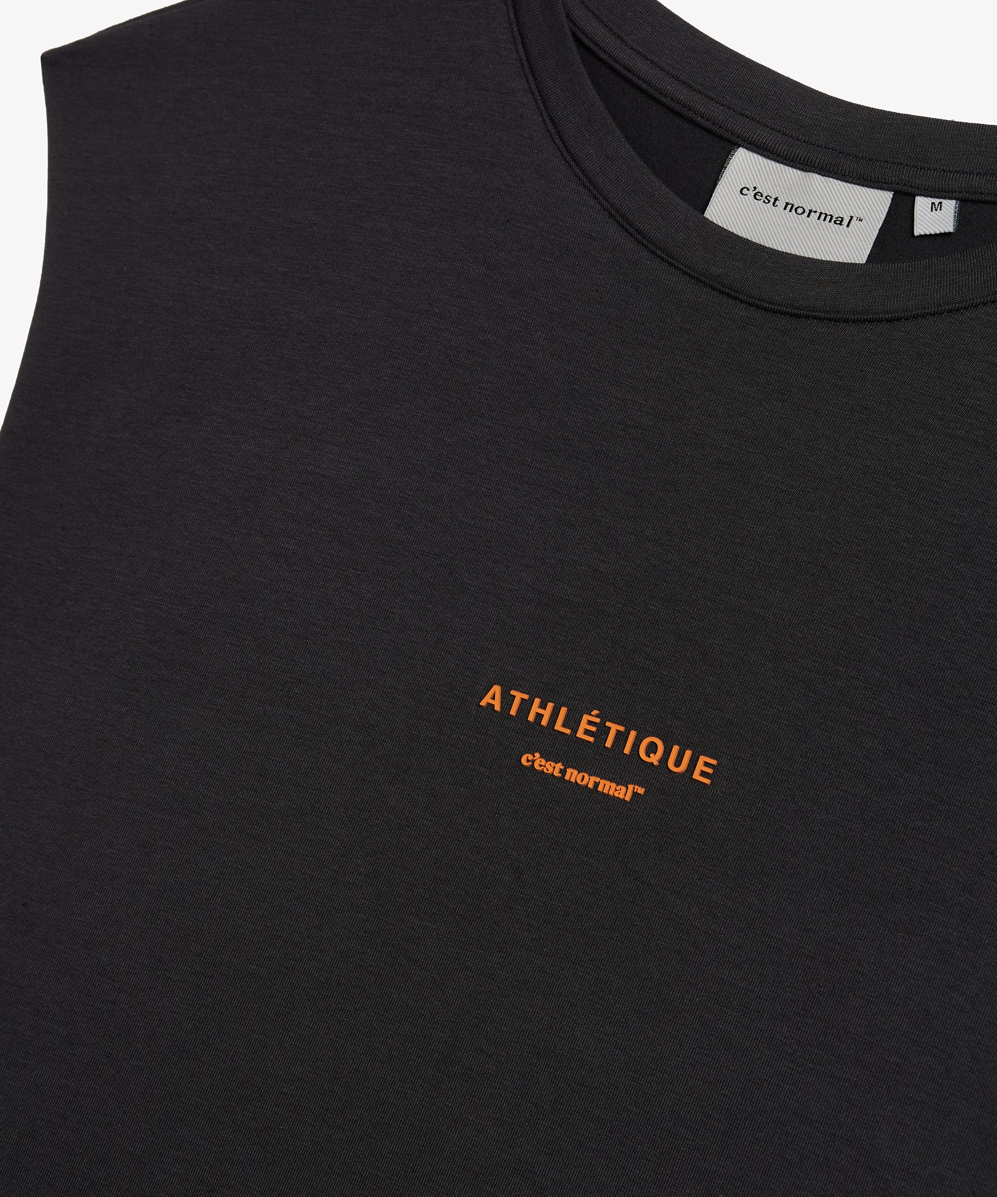 A detailed close-up of The Athlétique Tank in asphalt, focusing on the chest area. The image highlights the clean crewneck, the orange Athlétique and c'est normal logos printed on the front, and the inner neck label.