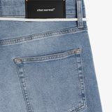 A close-up shot of the back of The Jeans Relaxed Straight Fit in Light Midwash. The image highlights the right back pocket, subtle orange accent stitching, and the black rubber patch with the white c'est normal logo at the back waist.