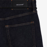 A close-up shot of the back of The Jeans Relaxed Straight Fit in Rinse Wash, laid flat on a white background. The image highlights the back pocket, subtle orange accent stitching, and the black rubber patch at the back waist featuring the c'est normal logo.