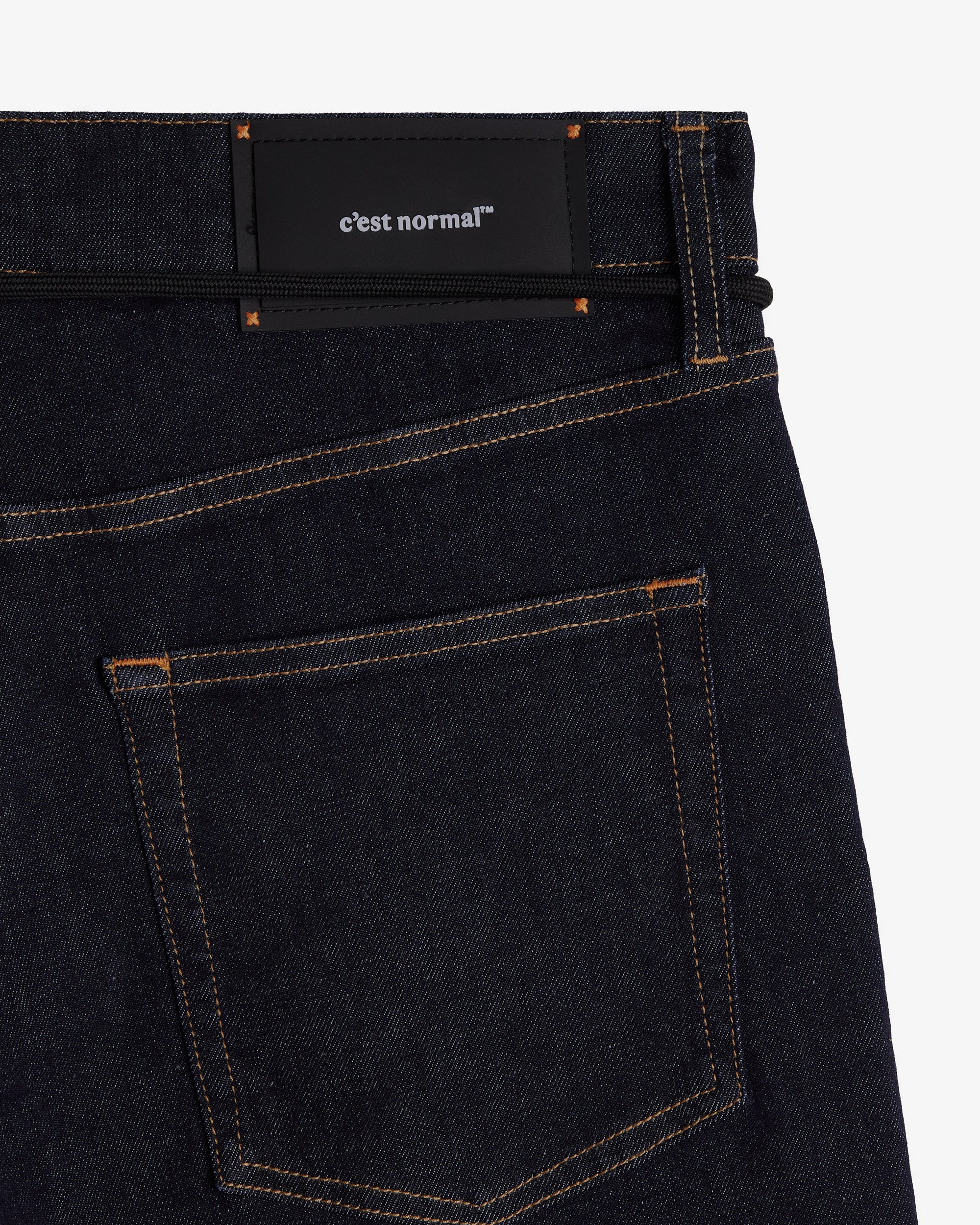 A close-up shot of the back of The Jeans Relaxed Straight Fit in Rinse Wash, laid flat on a white background. The image highlights the back pocket, subtle orange accent stitching, and the black rubber patch at the back waist featuring the c'est normal logo.