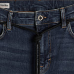 A close-up shot of the front waist area of The Jeans Skinny Fit in Midwash, highlighting the metal button, subtle orange accent stitching, and a black drawstring detail. A small white label with the c'est normal logo is visible on the inner waistband.