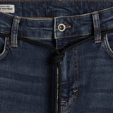 A close-up shot of the front waist area of The Jeans Skinny Fit in Midwash, highlighting the metal button, subtle orange accent stitching, and a black drawstring detail. A small white label with the c'est normal logo is visible on the inner waistband.