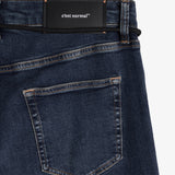 A detailed close-up of The Jeans Skinny Fit in midwash, laid flat on a white background. The image focuses on the back right pocket, the subtle orange accent stitching, and the black rubber patch at the waist featuring the white c'est normal logo.