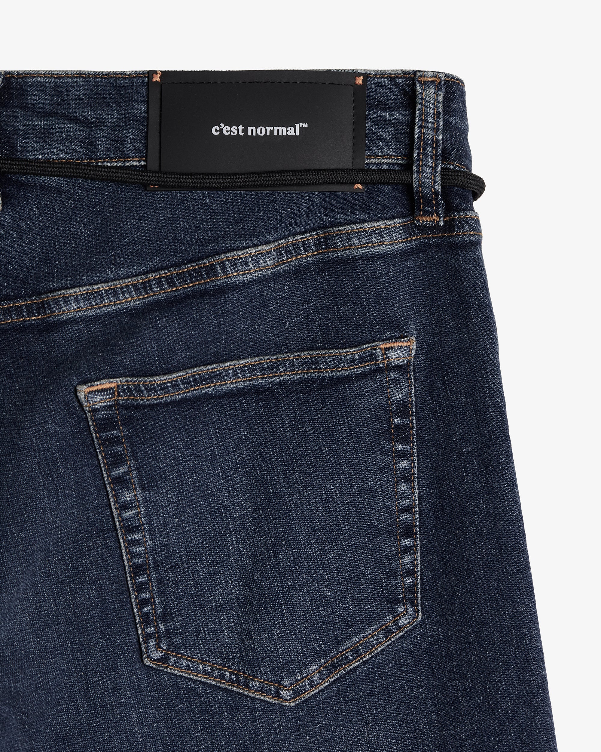 A detailed close-up of The Jeans Skinny Fit in midwash, laid flat on a white background. The image focuses on the back right pocket, the subtle orange accent stitching, and the black rubber patch at the waist featuring the white c'est normal logo.