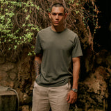 The Sanremo Linen Tee in mulled green — worn with beige trousers against a rocky wall