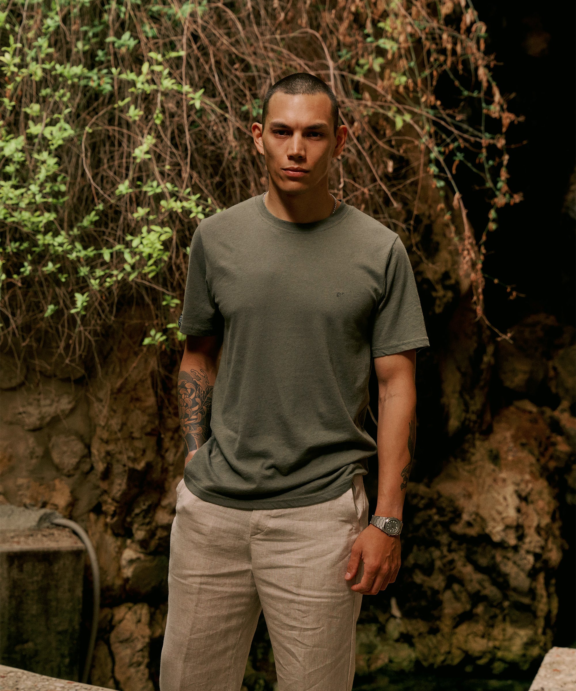 The Sanremo Linen Tee in mulled green — worn with beige trousers against a rocky wall