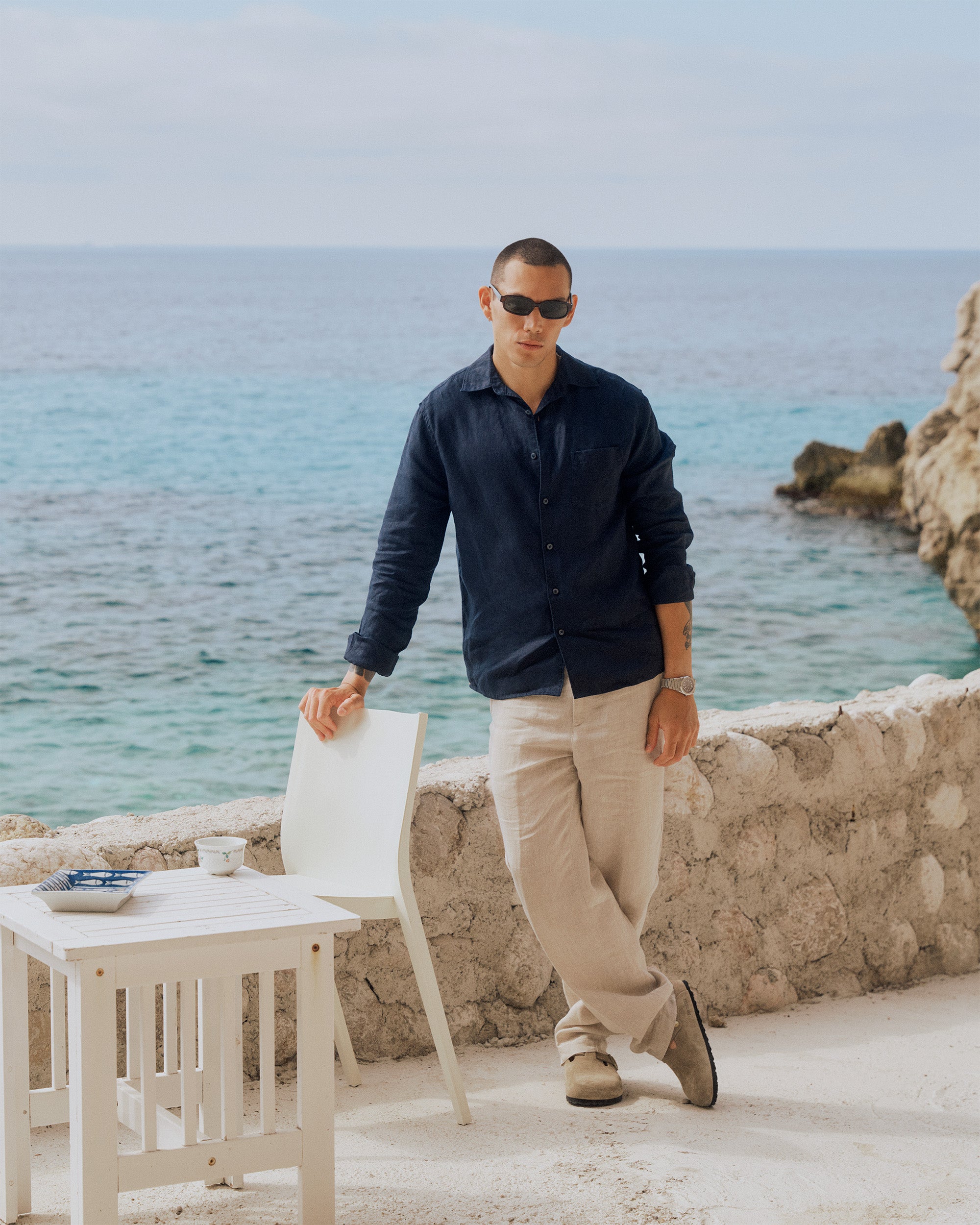 The Daily Linen Shirt in navy — worn with light trousers while standing by the sea