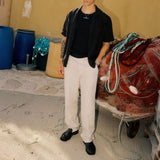 The Daily Linen Trouser in natural — worn with a black shirt and loafers near fishing nets